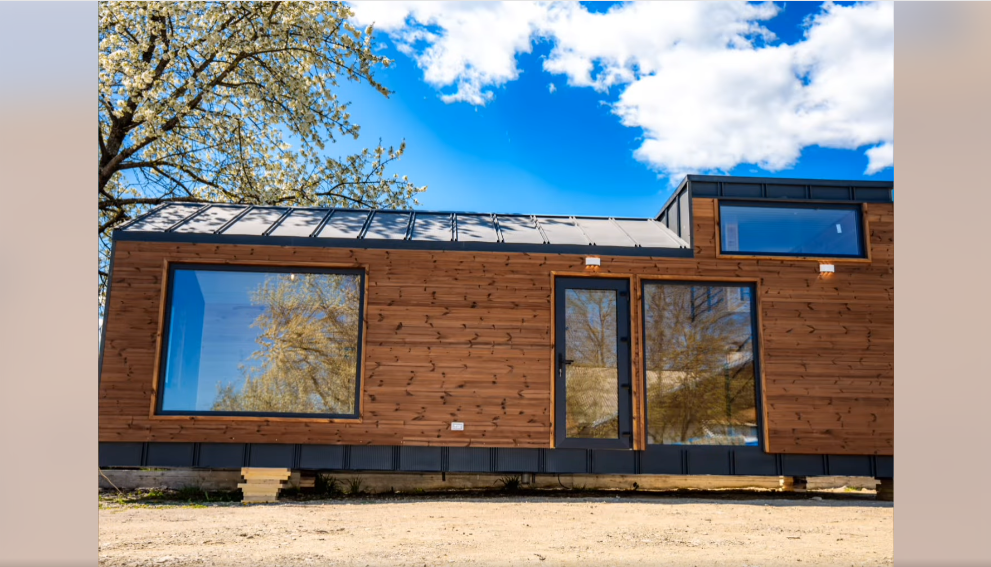 Lille træbeklædt tiny house med store vinduer og skråt tag mod blå himmel — Aktivus Tiny-kollektion.